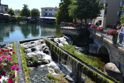 a river with a waterfall and a bridge and flowers at Appartement atypique dans un ancien couvent in LʼIsle-sur-la-Sorgue
